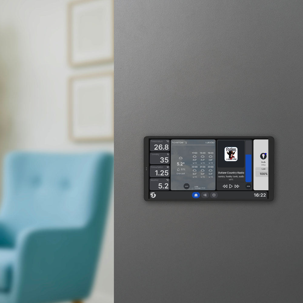 The Shelly Wall Display X2i Black is mounted on a gray wall, showing weather, temperature, radio, and other settings. In the background, a blurred blue armchair and framed pictures can be seen.